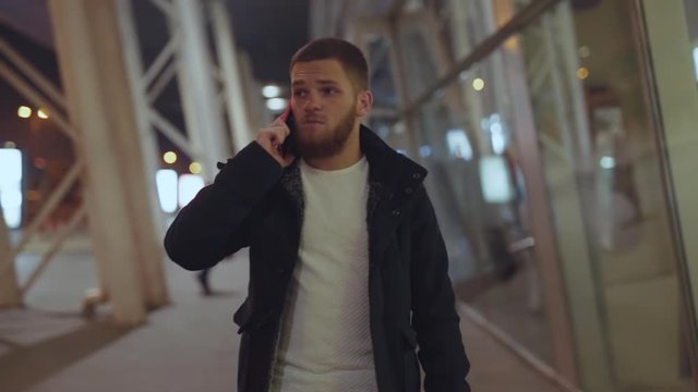 Portrait of handsome young man talking on mobile phone communicating walking on the street of modern city center during night time. Business guy. People and technology.