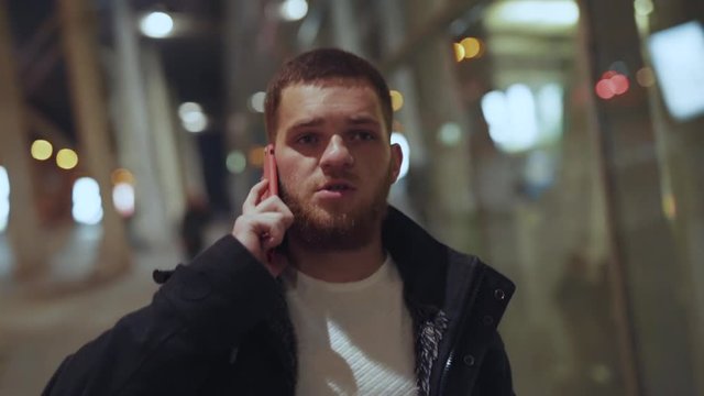Portrait of handsome young business guy talking on smartphone with partner walking serious in modern street of city center at night.