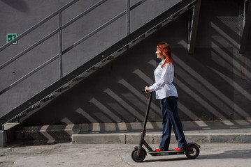 Fototapeta na wymiar A red-haired girl in a white shirt drives an electric scooter along the wall. A business woman in a trouser suit and red high heels rides around the city in a modern car. Business woman on a scooter.