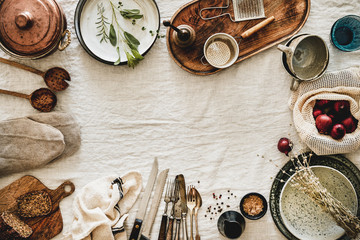 Flat-lay of various kitchen utensils, rustic tablewear, plates, dishes, glasswear, pan, mitten, textile for cooking over white linen tablecloth background, top view, copy space. Seasonal cooking