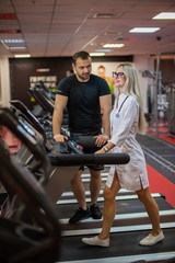 The athlete in the gym controls the treadmill exercises of a beautiful female doctor in medical uniform. The gym coach approves the slender figure of a beautiful physiotherapist woman.