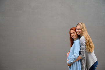 Same-sex relationships. A red-haired pregnant woman in a denim dress is standing against a gray wall, her hand is resting under her tummy. Her wife gently hugged the expectant mother from behind.