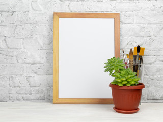 Empty wooden frame with white space for design. Frame, brushes and flower succulent in brown pot against white brick wall on white table.