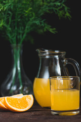 Freshly squeezed orange juice in a glass cup. Orange slices on a dark background. Glass vase with green branches.