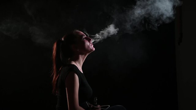 The brunette smokes an electronic cigarette on a black background, releases thick smoke from her mouth. Portrait of a woman smoking a vape, hovering.
