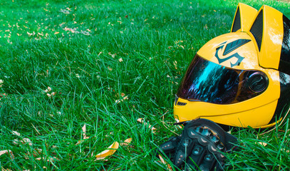 A yellow biker girl helmet from a motorcycle with mirror glass and ears and black biker gloves lies on green grass against the background of a gray motorcycle in autumn.