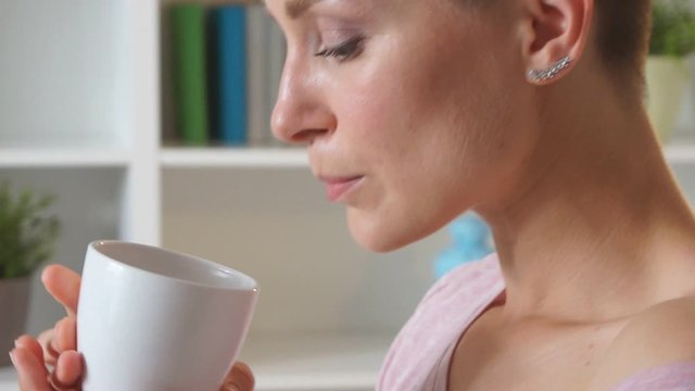 Young beautiful girl with short hair sit on bed in light room and enjoy drinking tea or coffee at morning at home.