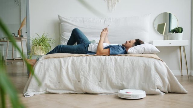 Guy is sufring the Internet with smartphone relaxing in bed while robotic vacuum cleaner is cleaning floor removing dust from laminate. Gadgets, people and housekeeping concept.