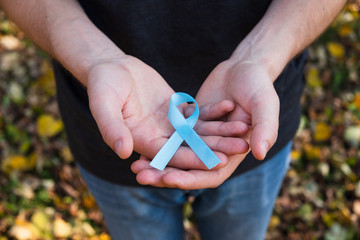 Man hands hold blue Prostate cancer awareness ribbon on fall foliage background. Medicine and...