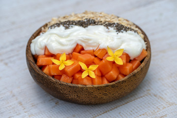 Slices of sweet papaya with oat flakes, chia seeds and white yogurt in coconut bowl on white wooden background, close up