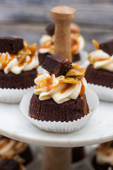 Chocolate cupcakes with cream cheese frosting, salted caramel sauce, brownie bites and toffees. Marble cakestand on grey wooden background.
