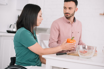young disabled woman holding bell pepper while talking to boyfriend in kitchen