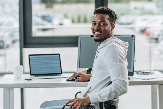 cheerful african american programmer smiling at camera while sitting at workplace in office