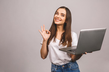 Portrait of a smiling young girl holding laptop computer and showing ok gesture isolated over grey...