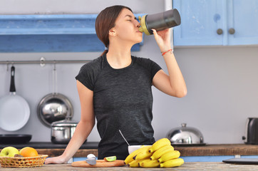Young woman drinking protein shake near table with ingredients in kitchen
