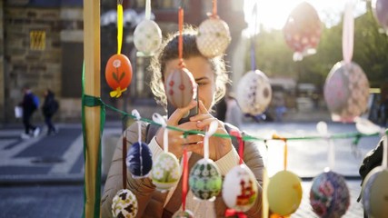 Woman tourist viewing gifts in street shop at famous easter market on old town square in Prague. Close-up portrait girl choosing painted easter egg hanging in sun shining, tradition religion symbol. 