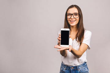 Fototapeta premium Nice idea! Surprised happy brunette woman showing blank smartphone screen while looking at the camera with open mouth over grey background.