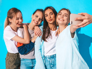 Four young smiling hipster women in summer clothes.Girls taking selfie self portrait photos on smartphone.Models posing near blue wall in studio,Female showing positive face emotions