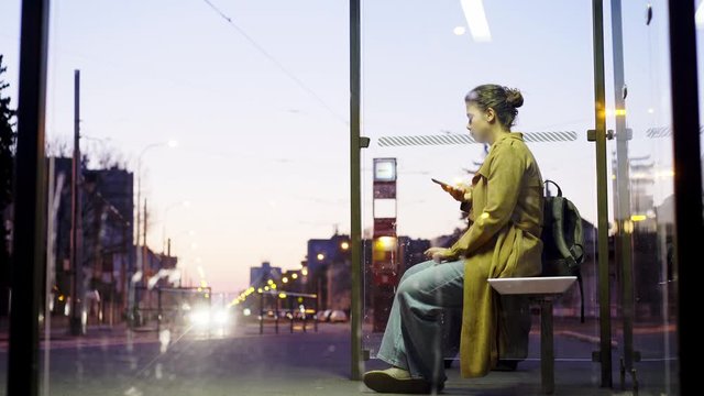 Illuminated bus stop in residential area on the outskirts of the city at night. View through glass of transparent shelter on roadside, sitting young girl using smartphone while waiting bus