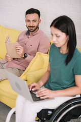 attentive disabled woman using laptop near smiling boyfriend sitting on sofa with book