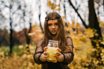 Pretty blonde girl in the autumn yellow fall park. Creative Halloween Day composition. Holiday celebration yellow candle skull glass in female hand with natural manicure.