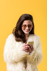 happy party girl in faux fur jacket and sunglasses using smartphone isolated on yellow