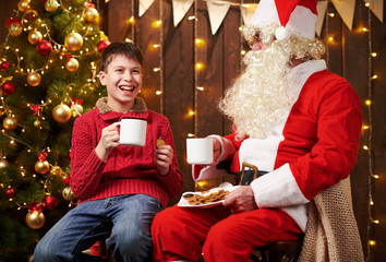 Santa Claus and child boy drinking tea, eating cookies, talking and sitting indoor near decorated xmas tree with lights - Merry Christmas and Happy Holidays!