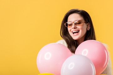 Fototapeta na wymiar happy party girl in faux fur jacket and sunglasses holding balloons isolated on yellow