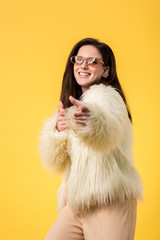 excited party girl in faux fur jacket and sunglasses pointing with finger at camera isolated on yellow