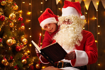 Fototapeta na wymiar Santa Claus and santa helper boy reading book, sitting indoor near decorated xmas tree with lights - Merry Christmas and Happy Holidays!