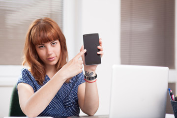 Naklejka premium Nice girl with tattoo and piercing showing a phone display (blank screen) and point at it with index finger, promoting app or website