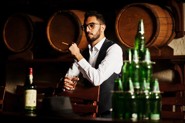 Handsome well-dressed arabian man with glass of whiskey and cigar posed at pub.