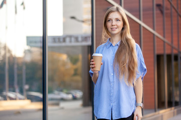 Happy smiling cute young woman with long hair holding a coffee in disposable paper cup, walking along the windows of office building.