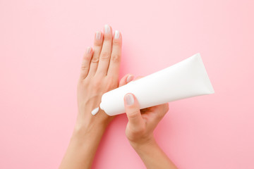 Young, perfect woman hands using moisturizing cream. Pastel pink background. Holding white tube. Care about clean, soft and smooth body skin. Point of view shot. - obrazy, fototapety, plakaty