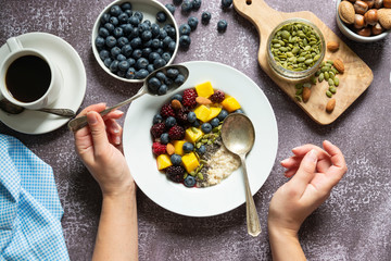 Healthy breakfast with oatmeal porridge, fruits and nuts. Blueberry, mango, pumpkin seeds, nuts, coffee cup. Top view with female hands. Clean eating, dieting, weight loss concept