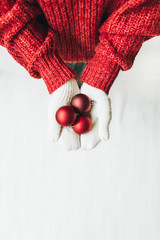 Christmas / New year / xmas composition. Young woman in red knitted sweater and white mittens holding tree red christmas toys. Flat lay, top view. Minimal winter holidays concept.