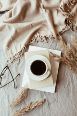 Autumn, fall composition. A cup of coffee lying on the grey linen bed with beige warm blanket, books, glasses and reeds. Lifestyle, hygge concept. Flat lay, top view.