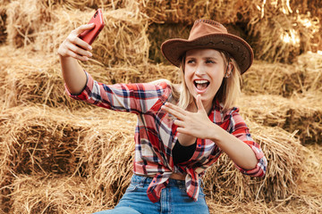Smiling young blonde cowgirl sitting on a haystack at the barn