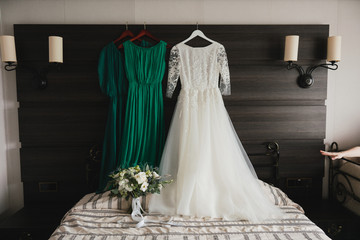 Modern bridal fashion. White wedding dress and green bridesmaids dresses hanging in the hotel room.