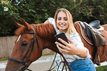 Foto op Canvas Beautiful smiling young blonde woman petting a horse at the stable © Drobot Dean