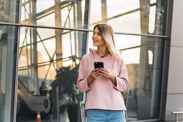 Young blonde female tourist in casual wear using smartphone while standing at airport with
