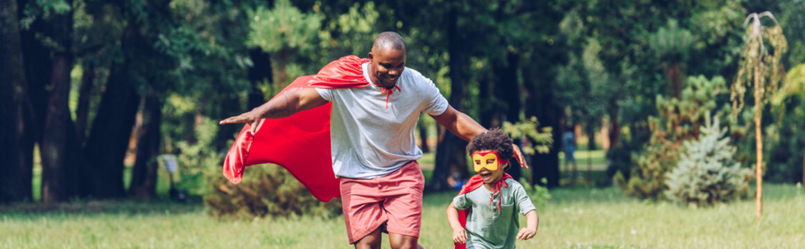 panoramic shot of african american father and son running in costumes of superheroes in park