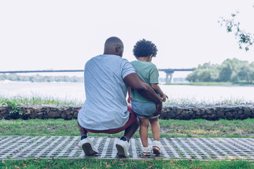 back view of african american father and son squatting and looking at river in park