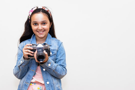 Teen girl dressed in a denim jacket smiling with a vintage camera in hands. There is a white background