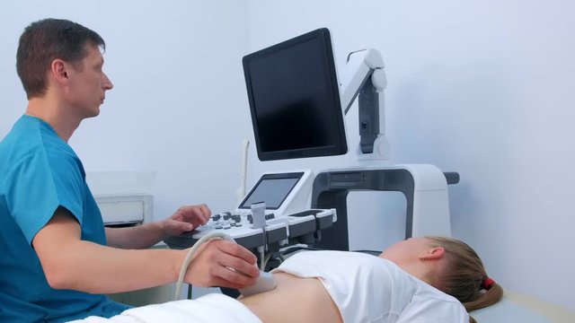 Man doctor examines abdominal ultrasound to diagnosis stomach of woman in clinic. He runs ultrasound sensor over patient's tummy and looks at image on screen. Diagnosis of internal organs.