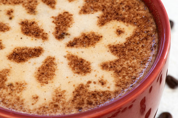 A foamy mug of cappuccino coffee with chocolate snowflake motif and a snowy background with a scattering of coffee beans sets the scene for a festive Christmas and warming up in cold winter weather