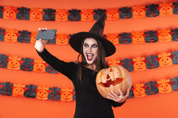 Image of witch girl in halloween costume taking selfie with pumpkin