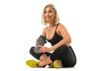 A sports girl with blonde hair and bright manicure in headphones, black sports topics, leggings and yellow sneaker holds a sports water bottle.