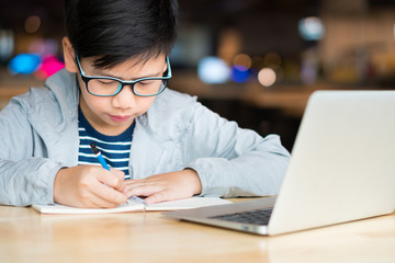 Smart looking Asian preteen boy writing and using computer laptop to study online lessons, research...