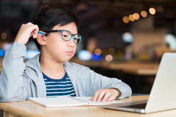 Smart looking Asian preteen boy holding pencil against his head, thinking and looking at computer laptop. Researching, studying and solving problem with concentration. Online learning concept.